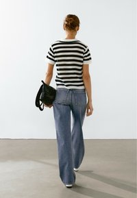 Haut en tricot à manches courtes rayé noir et blanc, associé à un jean bleu à jambe large. Sac à main texturé noir tenant dans la main droite.