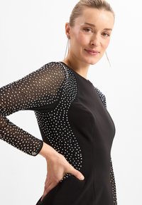 Robe noire avec des manches en tulle à pois transparentes ornées de bijoux argentés. Tissu lisse, design ajusté, encolure ronde et détails de silhouette subtils.