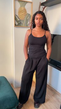 Black tank top and loose black trousers, with a smooth texture. Hair is wavy, and the background features a pineapple illustration.