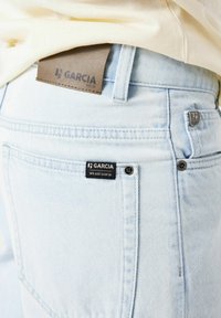 Los vaqueros de denim azul claro presentan una etiqueta de piel marrón con "GARCIA EST.72" y una etiqueta negra que dice "WE ARE GARCIA" en el bolsillo trasero.