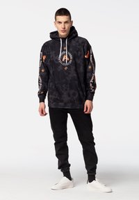 Crni hoodie s tie-dye uzorkom i bijelim vezicama, s šarenim grafičkim printovima na prednjoj strani i rukavima. Uparen s crnim hlačama i bijelim tenisicama.