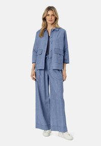 Giacca di jeans azzurro chiaro con maniche a tre quarti, bottoni frontali e due tasche a toppa, abbinata a pantaloni larghi coordinati e sneaker bianche.