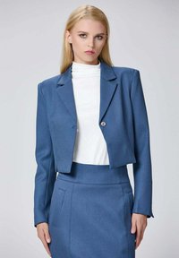 Blazer bleu cropped avec un seul bouton, tissu texturé, revers crantés et manches longues, porté sur un haut blanc à col montant.