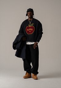 Sweatshirt preta com gráfico de maçã vermelha, jeans de ganga, botas castanhas e chapéu de balde preto. Segurando um casaco escuro, joalharia prateada visível.