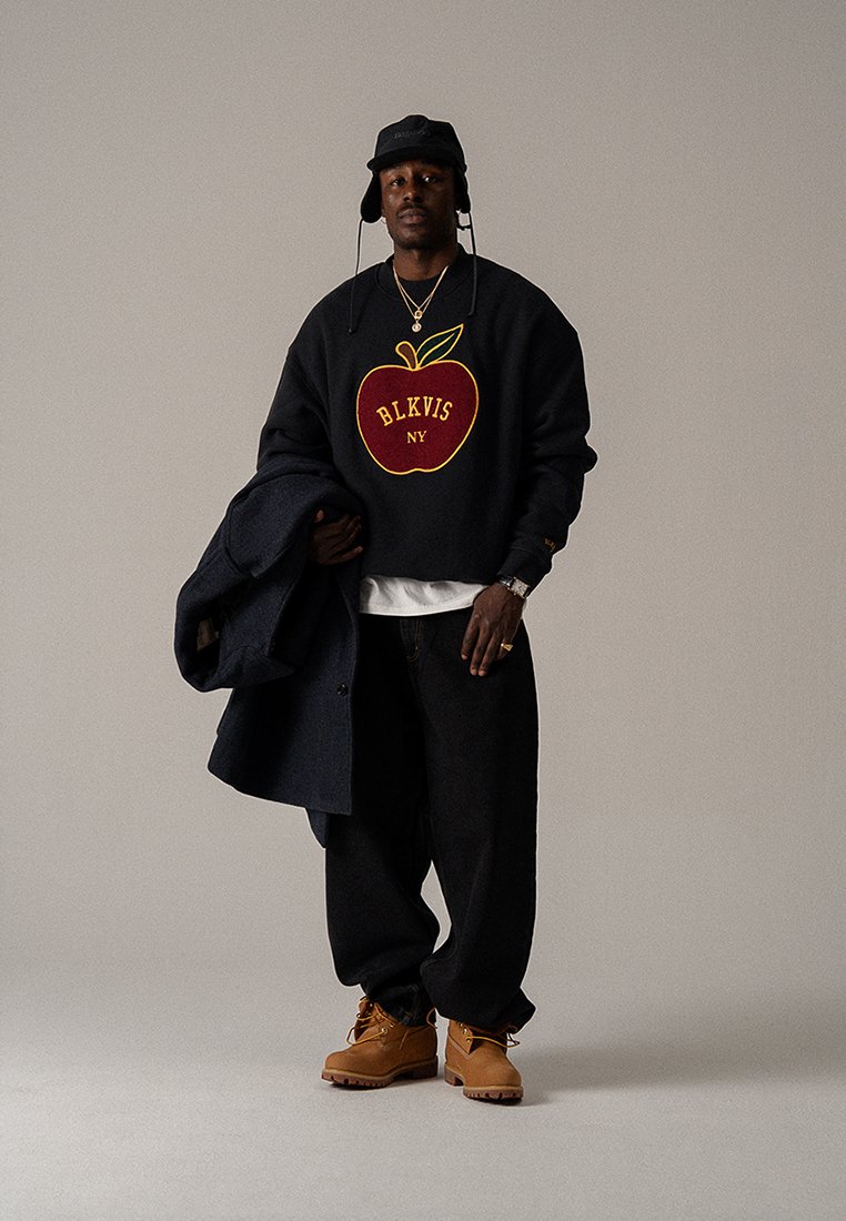 Sweatshirt preta com gráfico de maçã vermelha, jeans de ganga, botas castanhas e chapéu de balde preto. Segurando um casaco escuro, joalharia prateada visível.