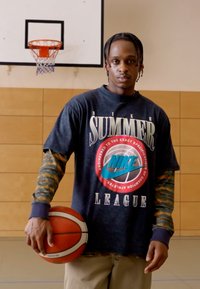 Nike Summer League t-skjorte, svart med fargerik logo, langermede grå og oransje mønstrede ermer, holder basketball i en gymsal.