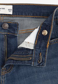 Μπλε τζιν παντελόνι με ανοιχτό φερμουάρ και κουμπί, ορατή εσωτερική ετικέτα που γράφει "made in Kenya" και μεταλλικό κουμπί μάρκας στη μέση.