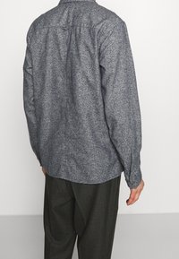 Mężczyzna w szarej teksturowanej koszuli z długim rękawem i ciemnych spodniach z wzorem, stojący tyłem do kamery na jednolitym tle.