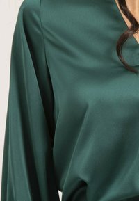 Blouse en soie vert foncé au toucher lisse, avec un col en V et des manches longues et fluides.