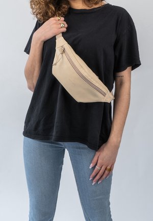 Person iført lyseblå jeans, sort T-shirt og beige crossbody taske med lynlås, holder taske strop med venstre hånd.