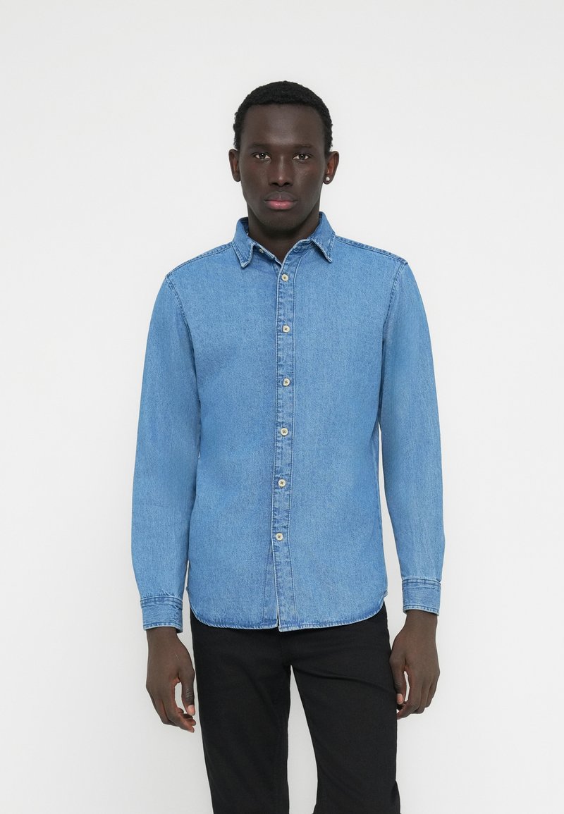 Hellblaues Jeanshemd mit langen Ärmeln, Knopfkragen, durchgehender Knopfleiste und kontrastierenden beigen Knöpfen. Kombiniert mit schwarzer Hose.