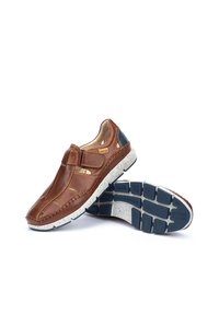 Zapatos de cuero marrón con un diseño perforado, tira de velcro y talón en contraste de color azul marino. Suela texturizada con una media suela blanca moteada.