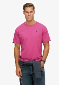 Camiseta de manga corta en algodón de color rosa brillante con cuello redondo y un pequeño logo negro en el pecho; combinada con jeans oscuros.