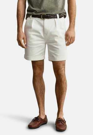 Mann trägt weiße, umgeschlagene Shorts, braunen gewebten Gürtel, olivgrünes T-Shirt und braune Lederloafer, steht vor einem einfarbigen Hintergrund.