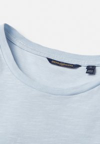 Maglietta di cotone blu chiaro con colletto a girocollo a costine e una texture sottile. L'etichetta mostra il nome del marchio "Henri Lloyd" e la taglia "M."