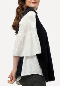 Top senza maniche in maglia blu navy sovrapposto a una blusa bianca con texture e maniche a campana, indossato da un modello visto dal profilo.