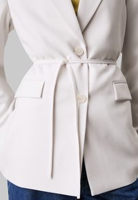 Biały żakiet o dopasowanym kroju z paskiem w talii, z pojedynczym zapięciem na guzik i naszytymi kieszeniami. Gładka tekstura i długie rękawy.