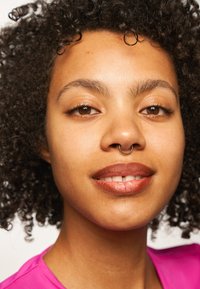 Nærbilde av en person med krøllete hår, iført en rosa topp. Kjennetegn inkluderer en nesering og naturlig sminke med brune toner.