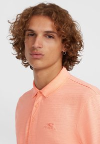 Polo shirt en corail clair avec un tissu texturé, doté d'un col, de manches courtes et d'un logo brodé sur la poitrine.