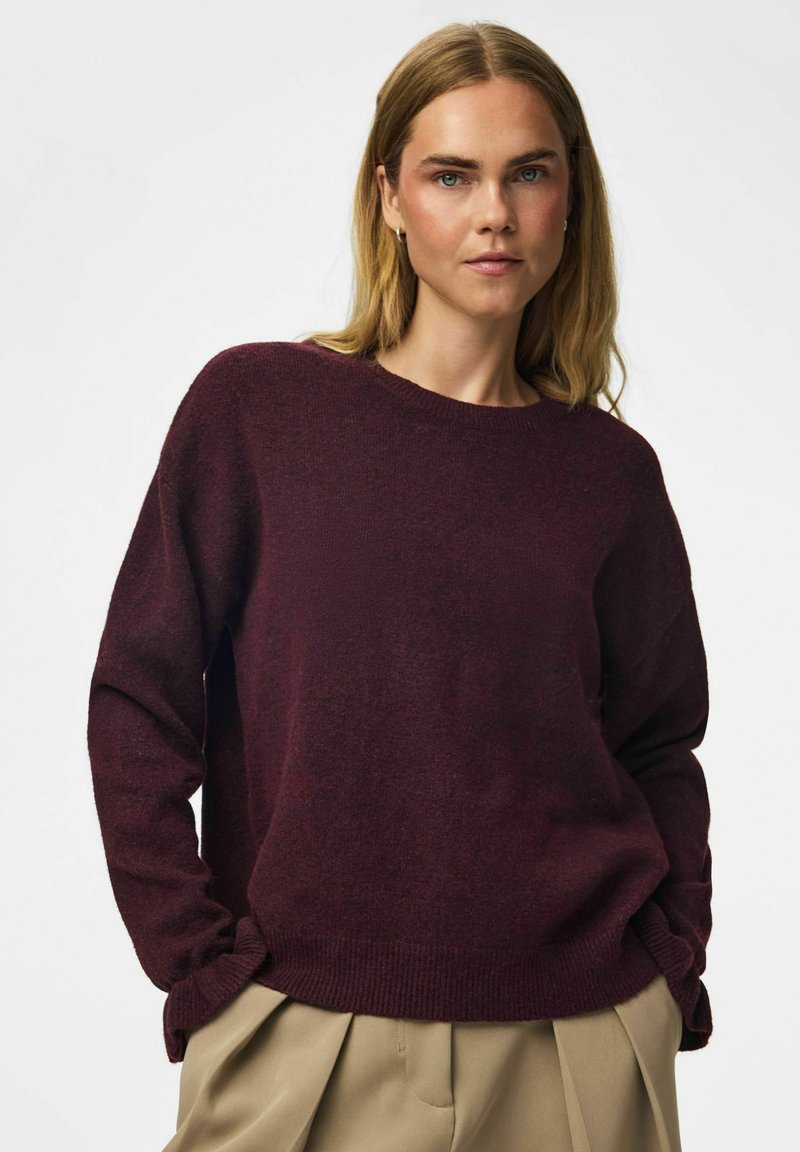 Femme blonde à la peau claire portant un pull bordeaux foncé et un pantalon plissé beige, debout les mains dans les poches devant un fond blanc.