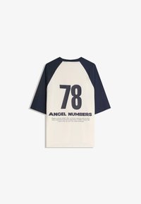 Bela majica s mornarsko modrimi raglan rokavi, z velikim napisom "78" in besedilom "ANGEL NUMBERS" na hrbtu, ter dodatnimi natisnjenimi podrobnostmi spodaj.