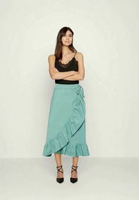 Uma saia envolvente azul-água com uma bainha em babados e detalhe de amarração lateral, combinada com uma blusa preta com rendas e saltos pretos com tiras.