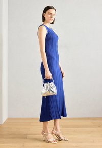 Robe midi en tricot bleu avec des plis verticaux, un col rond et des accents de boutons dorés. Un sac à main avec cordon en argent et des sandales beiges à lanières complètent le look.