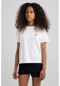 Weißes Baumwoll-T-Shirt mit kurzen Ärmeln und Rundhalsausschnitt, mit einem roten Grafikedruck auf der linken Brustseite. Kombiniert mit schwarzen Shorts.