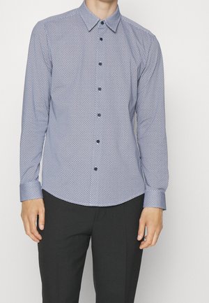 Man draagt een nauwsluitend, patroonlang mouw overhemd met zwarte broek, staand tegen een effen witte achtergrond.