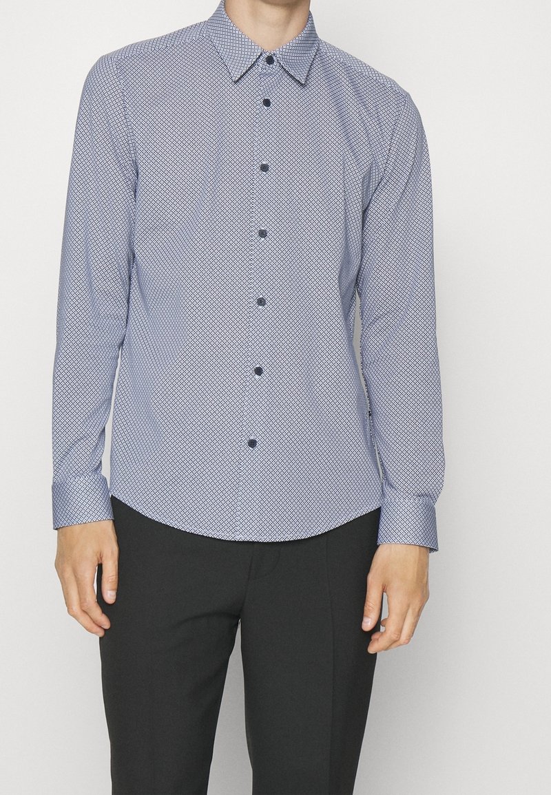 Homme portant une chemise habillée ajustée à manches longues avec motifs et un pantalon noir, debout devant un fond blanc uni.