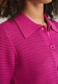Fuchsia stickad cardigan med ett texturerat mönster, knäppning fram och spetsig krage. Materialet är lätt och andas.