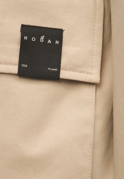 Μπεζ ύφασμα με μια μαύρη διπλωμένη ετικέτα που γράφει "HOGAN," "1986," και "MILANO," ραμμένη κοντά σε μια ραμμένη άκρη.