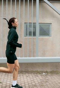 Mulher vestindo um traje atlético preto a correr junto a uma cerca de metal e uma parede bege com uma pequena janela ao ar livre.