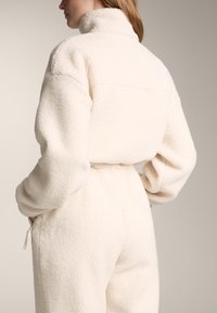 Flis jopi v barvi slonokoščene kosti, z visokim ovratnikom in teksturirano, puhasto tkanino. Vključuje šiv na hrbtni strani in oprijet pas.