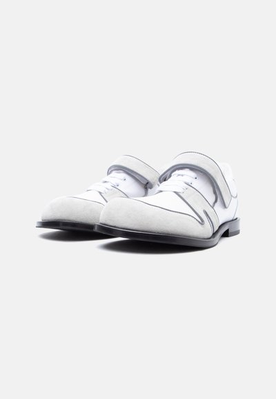 Ζευγάρι λευκά και γκρι παπούτσια φόρμας με κορδόνια και λουράκι βέλκρο, με μαύρες σόλες, τοποθετημένα δίπλα δίπλα σε απλό ανοιχτόφωτο φόντο.