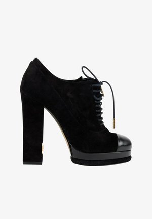Chaussure à talon haut en daim noir avec semelle plateforme, bout en cuir noir et lacets noirs avec embouts dorés, vue de profil.