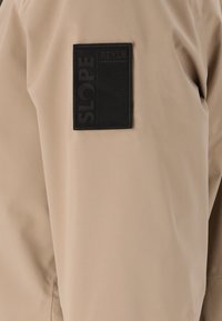 Beige jacka med en jämn, matt yta. Har en svart rektangulär lapptikett med texten "SLOPE STYLE" på vänster sida.