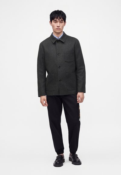 Jeune homme portant une veste à carreaux foncés boutonnée, un pantalon noir et des mocassins noirs, debout devant un fond blanc uni.