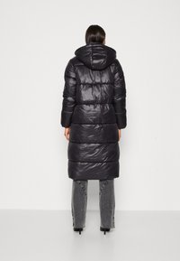 Manteau matelassé noir à capuche, avec une texture brillante et matelassée, une silhouette longue et de larges panneaux horizontaux. Porté avec un pantalon gris.