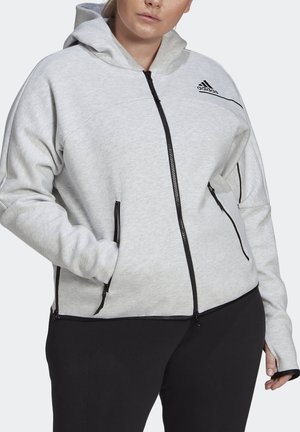 Woman wearing light gray Adidas hoodie with black zippers and logo, hands in pockets, paired with black pants.