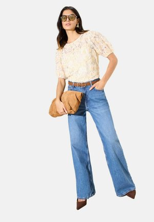 Nő áttetsző csipke krémszínű blúzban, magas derekú kék farmerral, barna övvel, barna hegyes orrú cipővel, feltűnő fülbevalókkal, napszemüvegben, kezében nagy barna borítéktáskával.