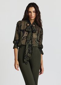 Blusa verde trasparente con motivo paisley nero, colletto a fiocco e maniche lunghe, abbinata a pantaloni aderenti color oliva.