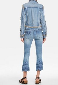 Chaqueta de mezclilla azul claro con bordes deshilachados, combinada con jeans claros ajustados que presentan patrones florales bordados en los tobillos. Sandalias de cuña.