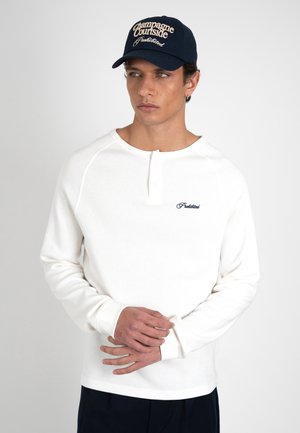 Mand iført hvid langærmet henleytrøje med lille "Forbudt" tekst, navy blå kasket med teksten "Champagne Courtside Prohibited", hænderne foldet.