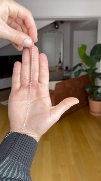 O mână ține o brățară din argint, întinsă vertical, pe un fundal de sufragerie cu o plantă și o canapea.