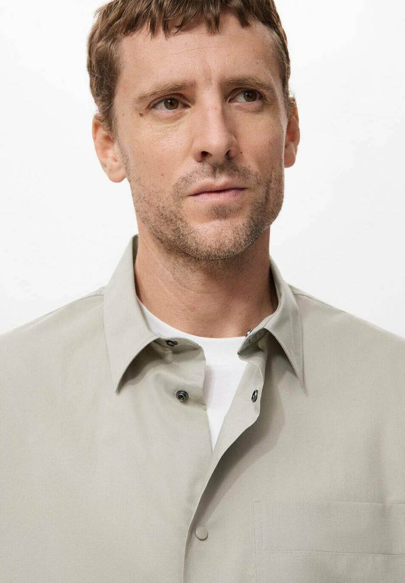 Mann mit kurzen braunen Haaren und leichten Bartstoppeln, der ein beiges, knopfbares Hemd über einem weißen T-Shirt trägt und leicht nach rechts schaut.