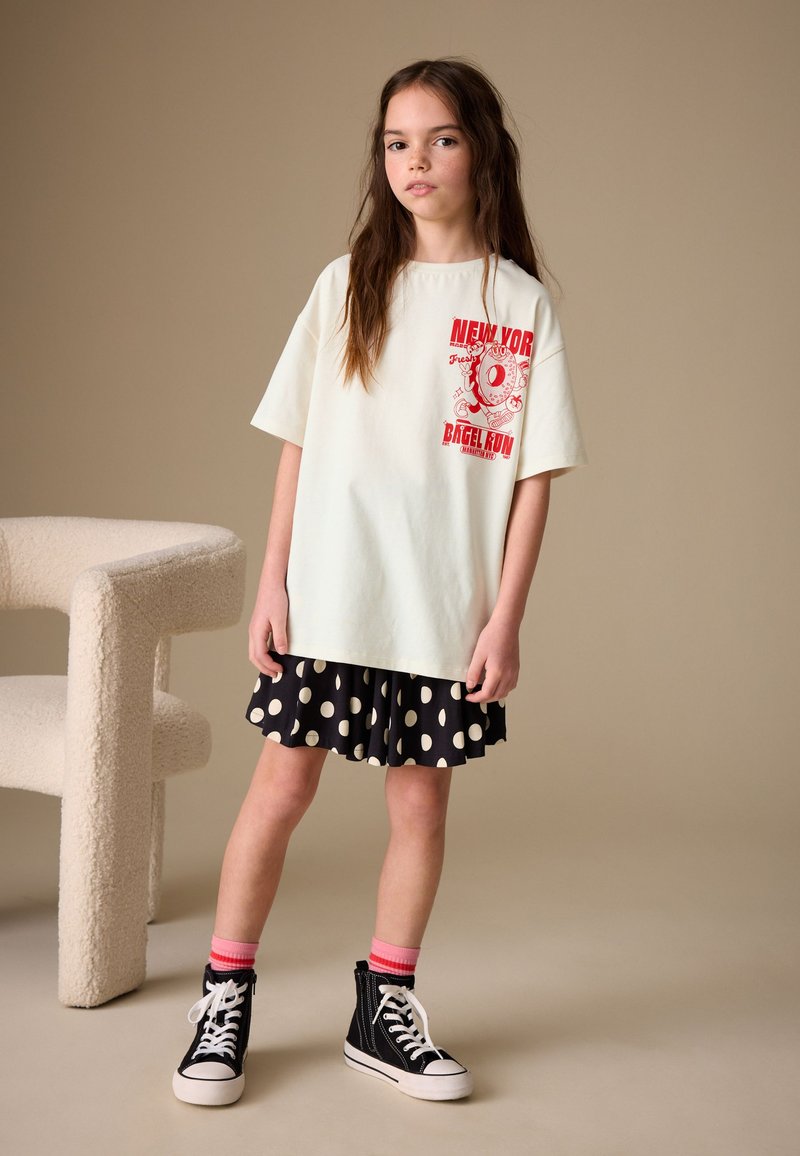 Jeune fille se tenant à côté d'une chaise beige texturée, portant un t-shirt blanc oversize avec un imprimé rouge, une jupe noire à pois, des chaussettes roses et des baskets noires.