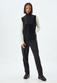 Sort sporty vest med lynlås fortil, fleece-krop og vandafvisende panel. Sammen med sorte tætsiddende bukser og sorte sportssko.