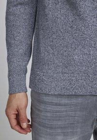 Gray textured sweater with ribbed cuffs and hem, worn over light gray pants. Visible hand resting at the side.