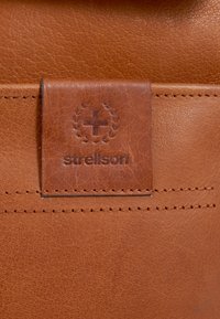 Hnědá kožená nášivka s embosovaným logem "strellson" a kruhovým emblémem, s kontrastním prošíváním na texturovaném pozadí.